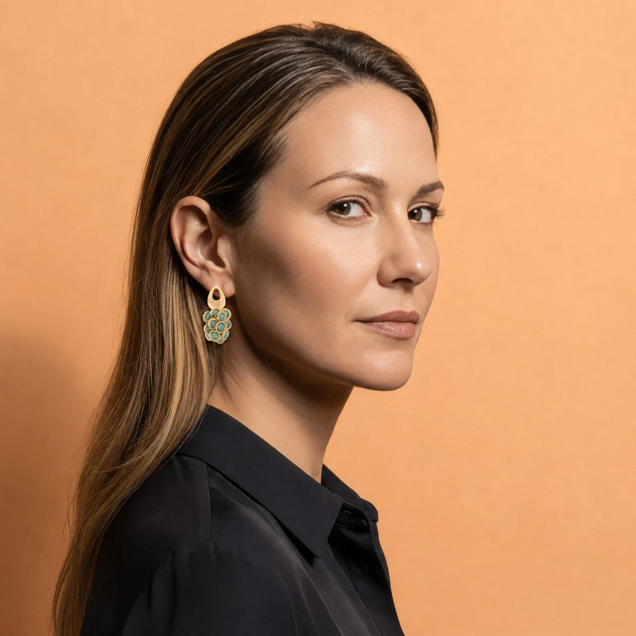 A woman in a black collared shirt wearing a gold and green drop earring, shown in profile against a solid peach background.