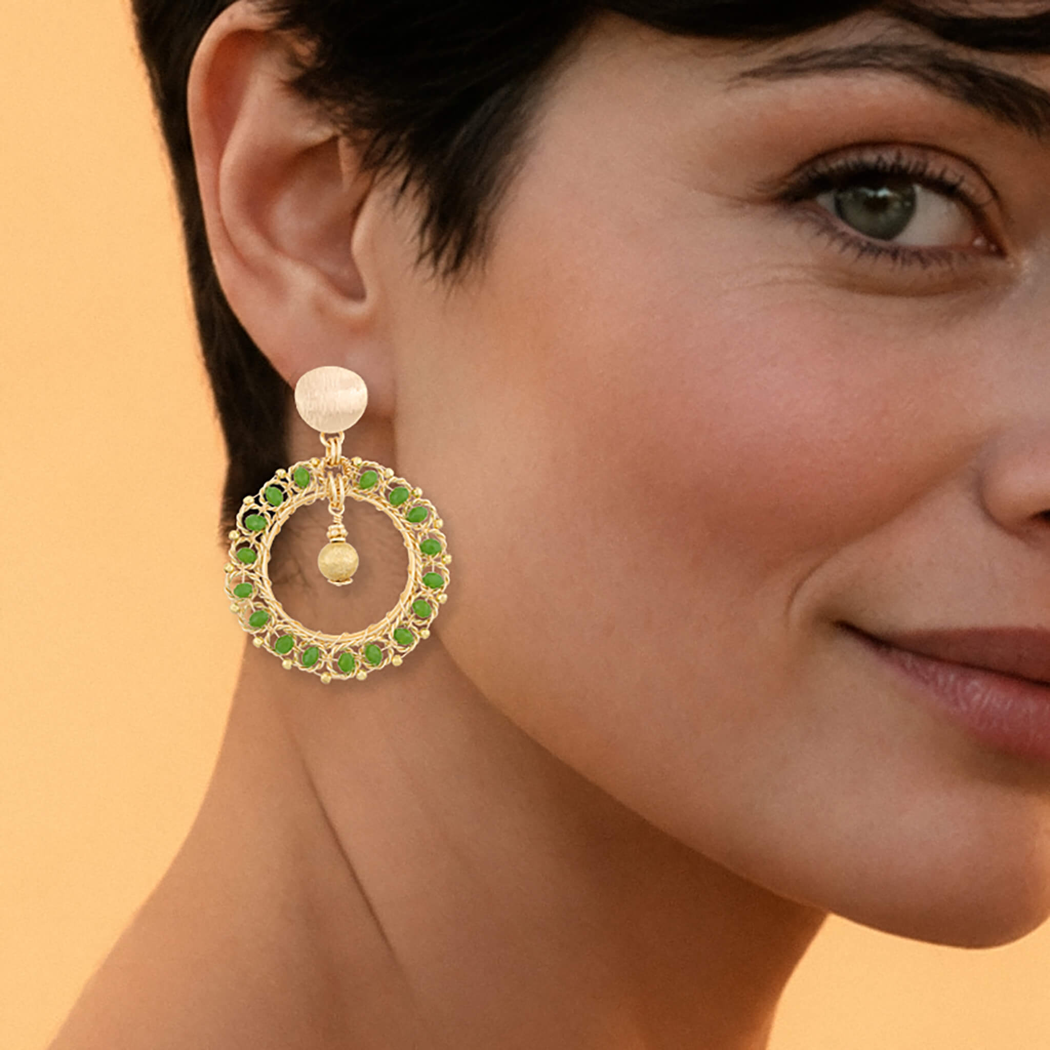 A close-up of a model's ear wearing a gold circular drop earring adorned with green Czech crystals and a central gold bead, set against a warm peach background.