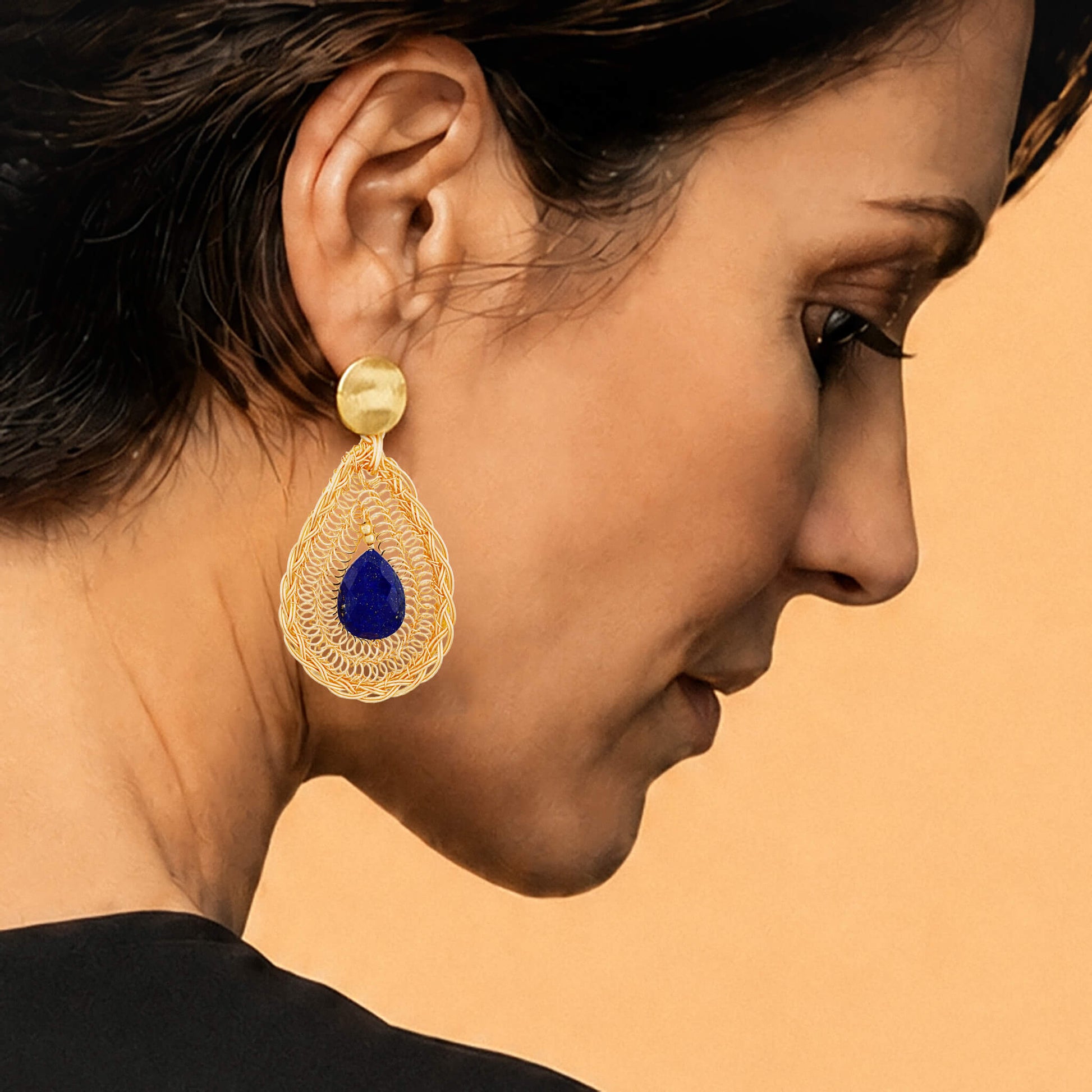 A close-up side profile of a model wearing a large gold teardrop earring with a deep blue Lapis Lazuli gemstone in the center, set against a solid warm beige background.