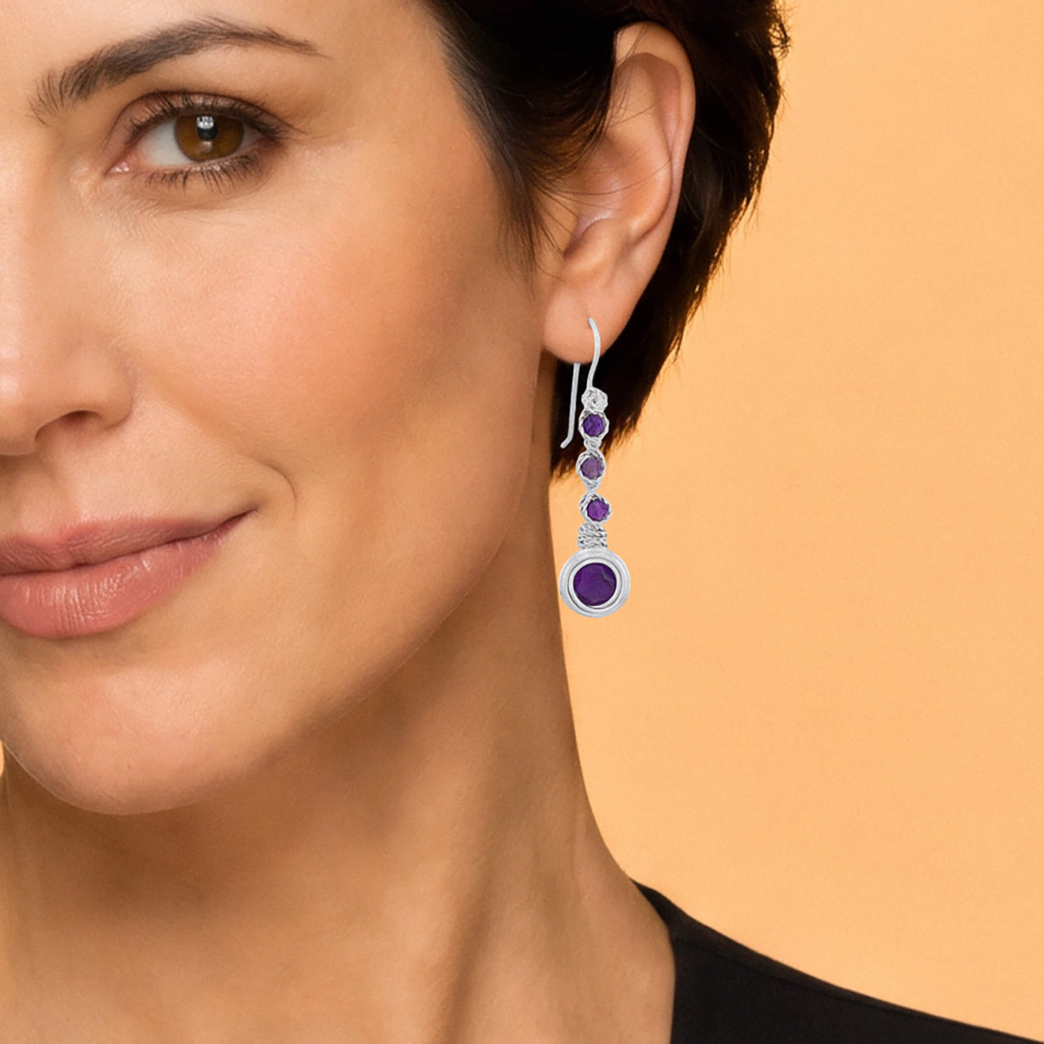 A close-up of a model wearing a silver drop earring with purple amethyst gemstones, set against a solid peach background.