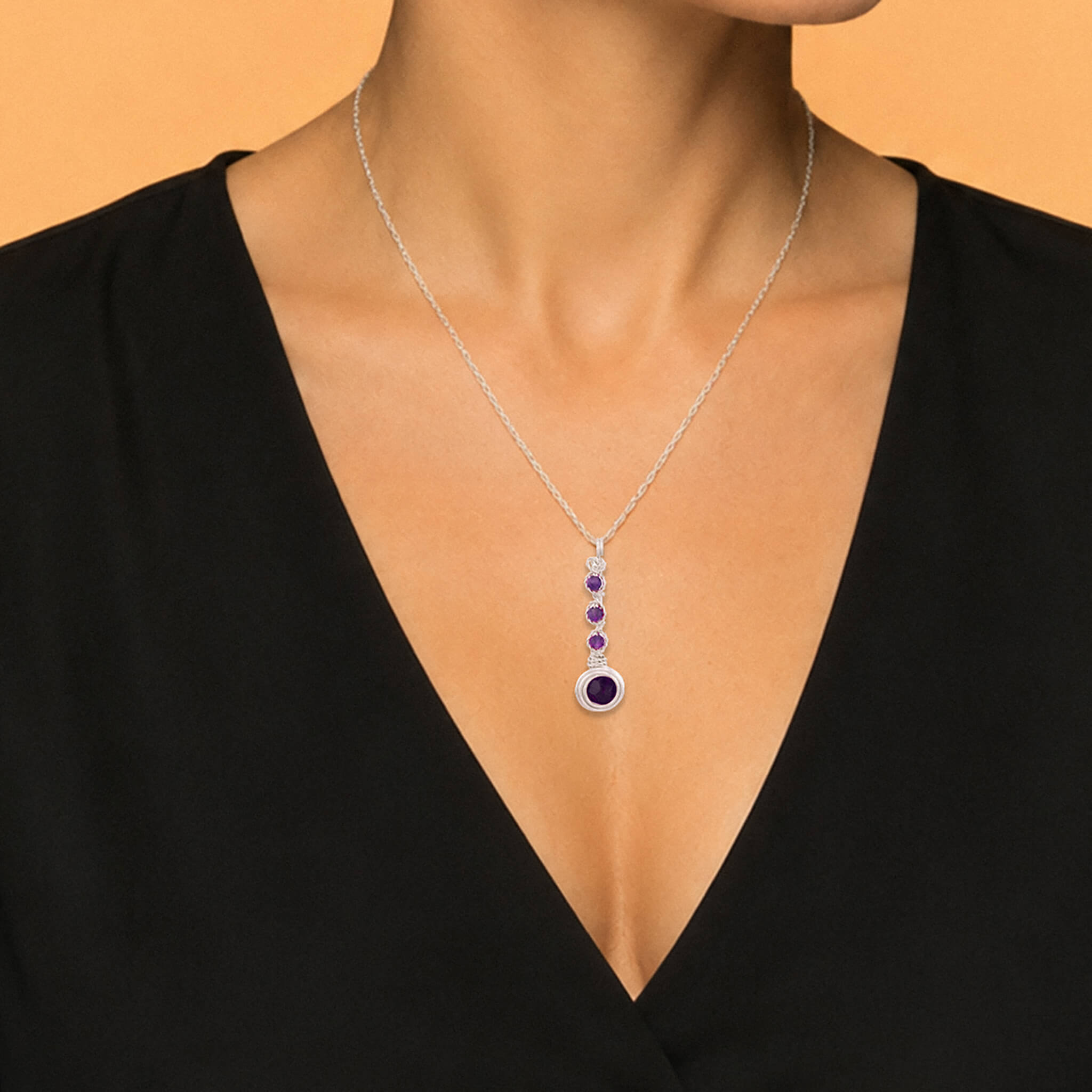 A silver pendant necklace with purple gemstones worn by a model in a black V-neck top against a warm beige background.