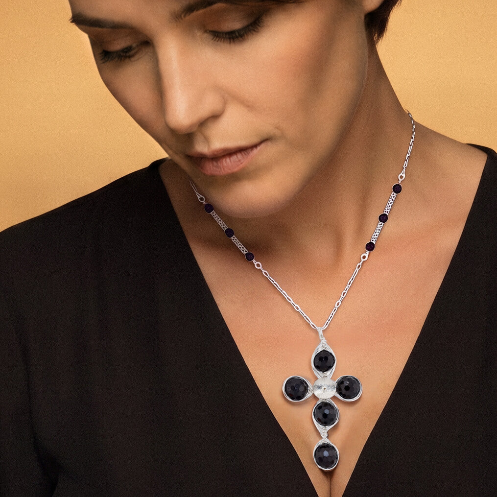 A close-up of a model wearing a silver cross pendant necklace with dark red garnet round faceted stones, set against a warm peach background.