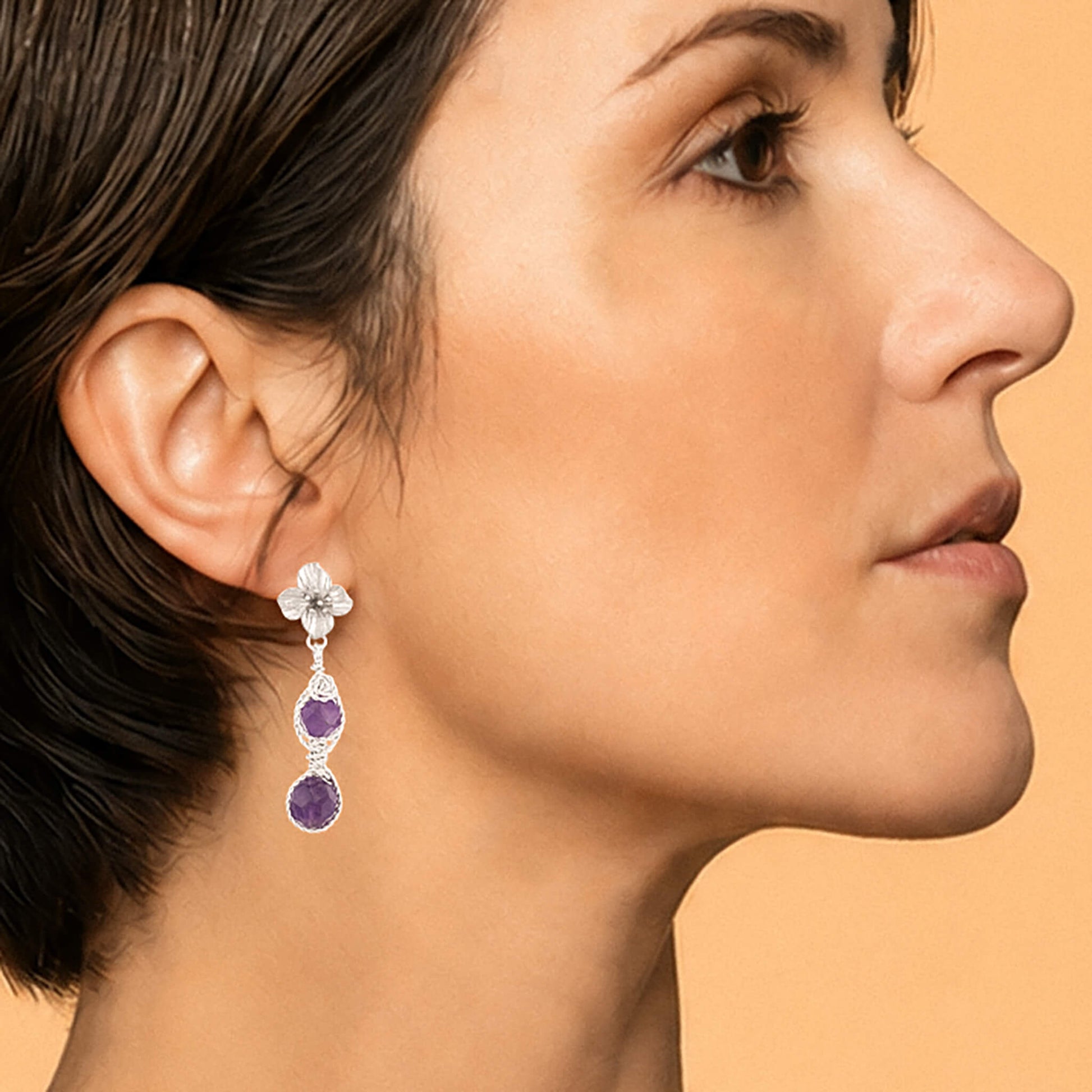 A close-up side profile of a woman wearing a dangling earring. The earring features a silver flower stud setting with small clear stones and two purple round amethyst stones below, set against a solid warm beige background.
