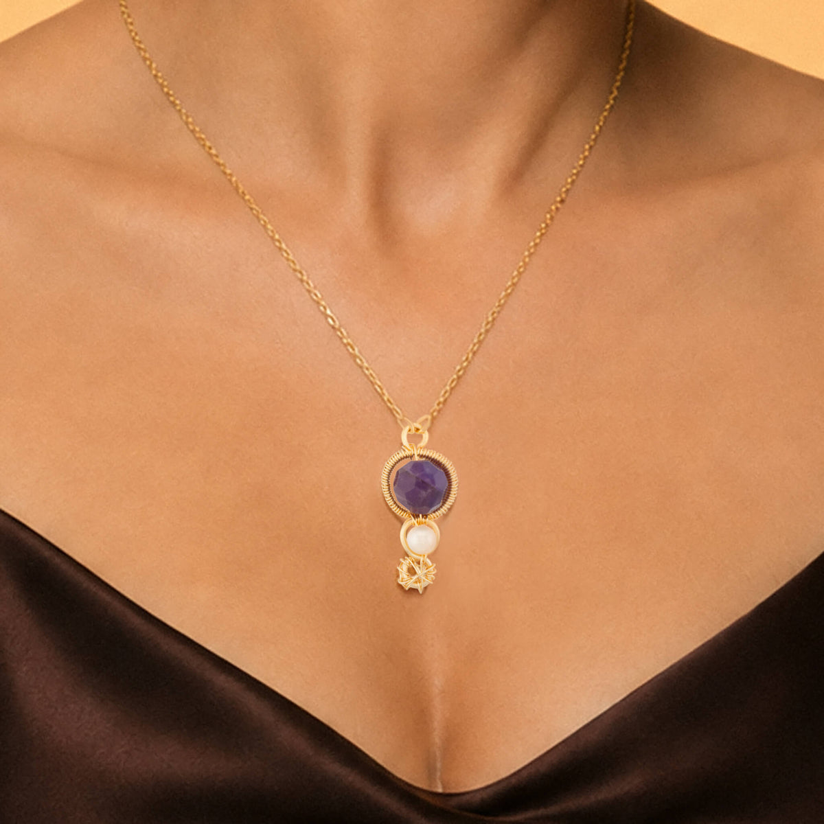 A gold chain necklace with a pendant featuring a purple round Amethyst gemstone, a white oval stone, and a small freshwater pearl, shown worn on a model's neck.