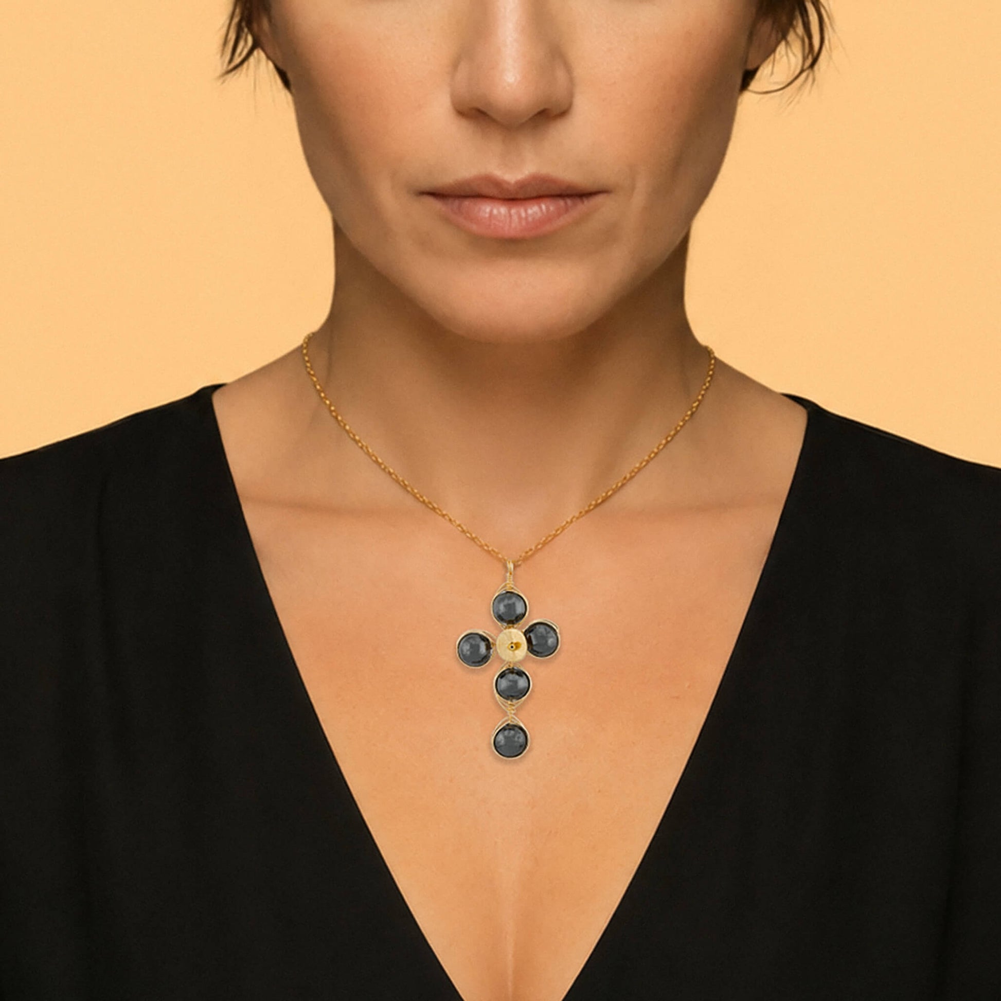 A close-up of a model wearing a gold cross pendant necklace with dark silver round Hematite gemstones, set against a solid warm beige background.