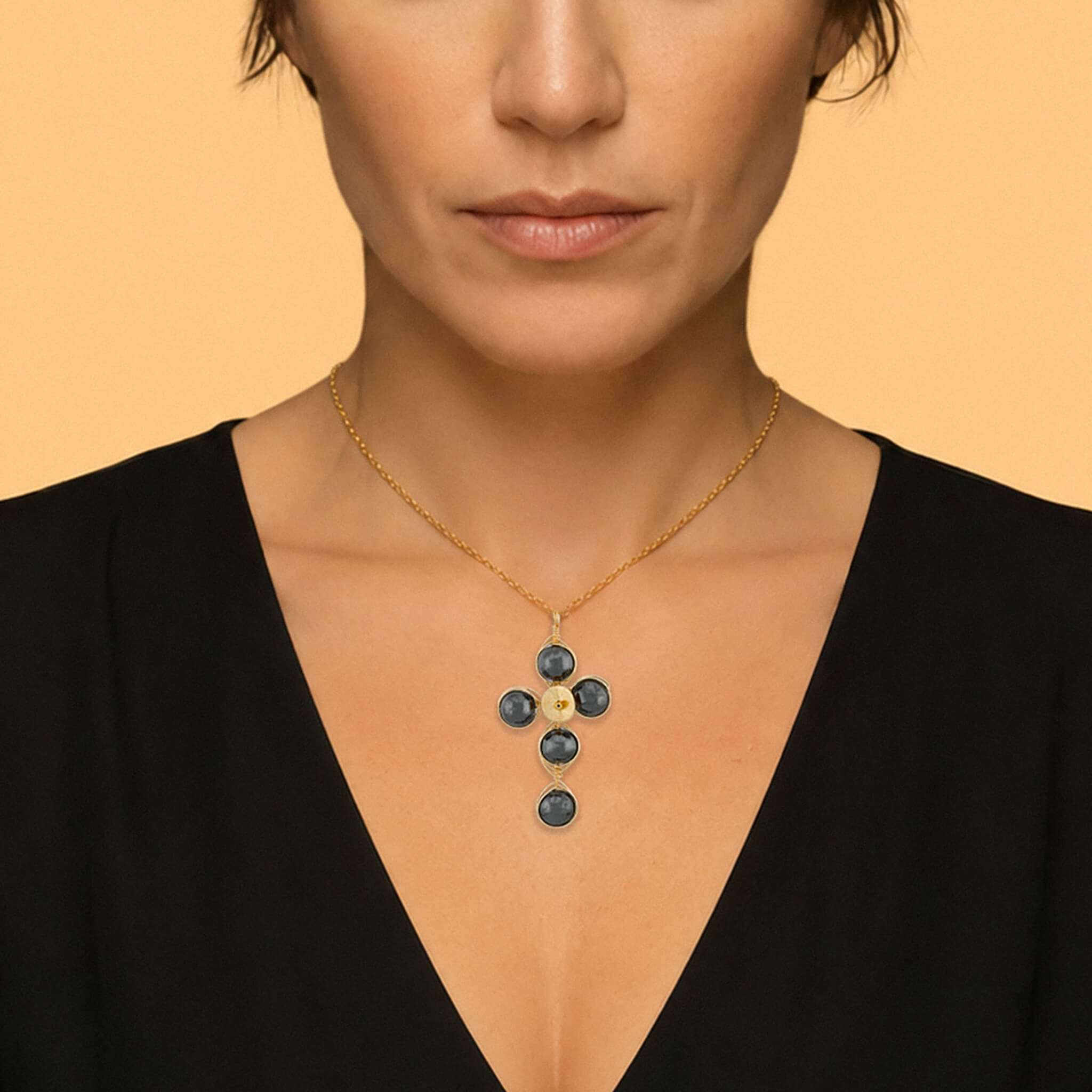 A close-up of a model wearing a gold cross pendant necklace with dark silver round Hematite gemstones, set against a solid warm beige background.