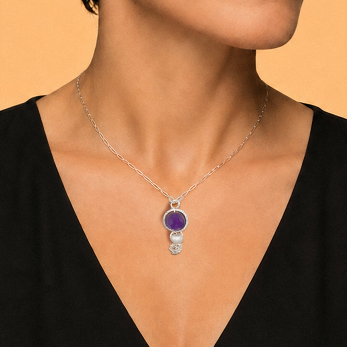 A close-up of a model wearing a silver chain necklace with a pendant featuring a purple round Amethyst gemstone and a small freshwater pearl below it, set against a warm beige background.