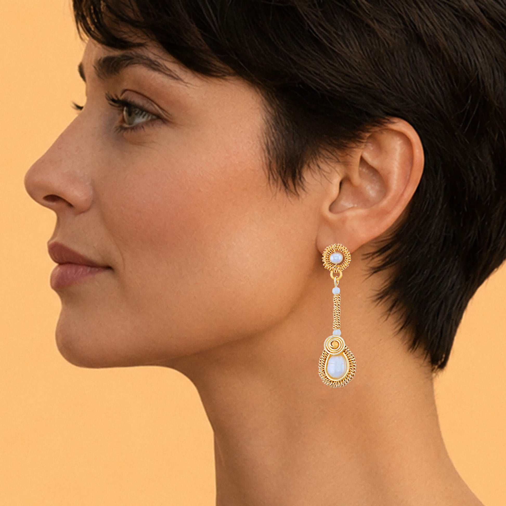 Close up of model wearing a gold dangle earring with freshwater pearls accents, set against a solid warm peach background.