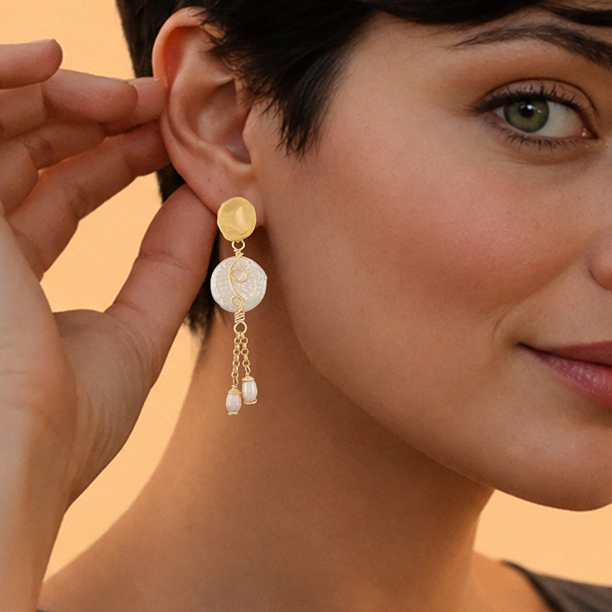 A close-up lifestyle shot of a woman wearing a gold and white dangle earring, with her hand adjusting the earring on her ear. The earring features a gold disc, a white textured disc, and two small dangling pearl-like beads.