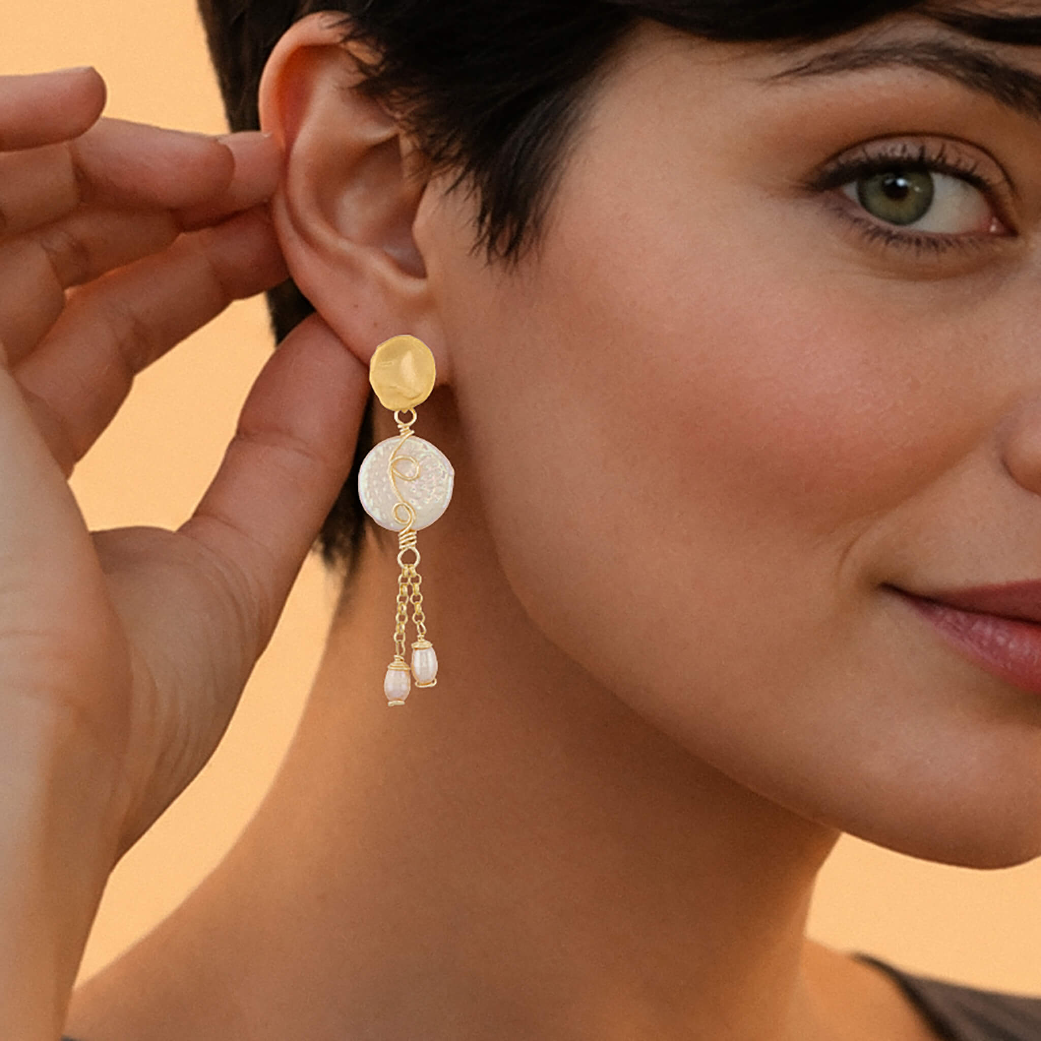 A close-up lifestyle shot of a woman wearing a gold and white dangle earring, with her hand adjusting the earring on her ear. The earring features a gold disc, a white textured disc, and two small dangling pearl-like beads.