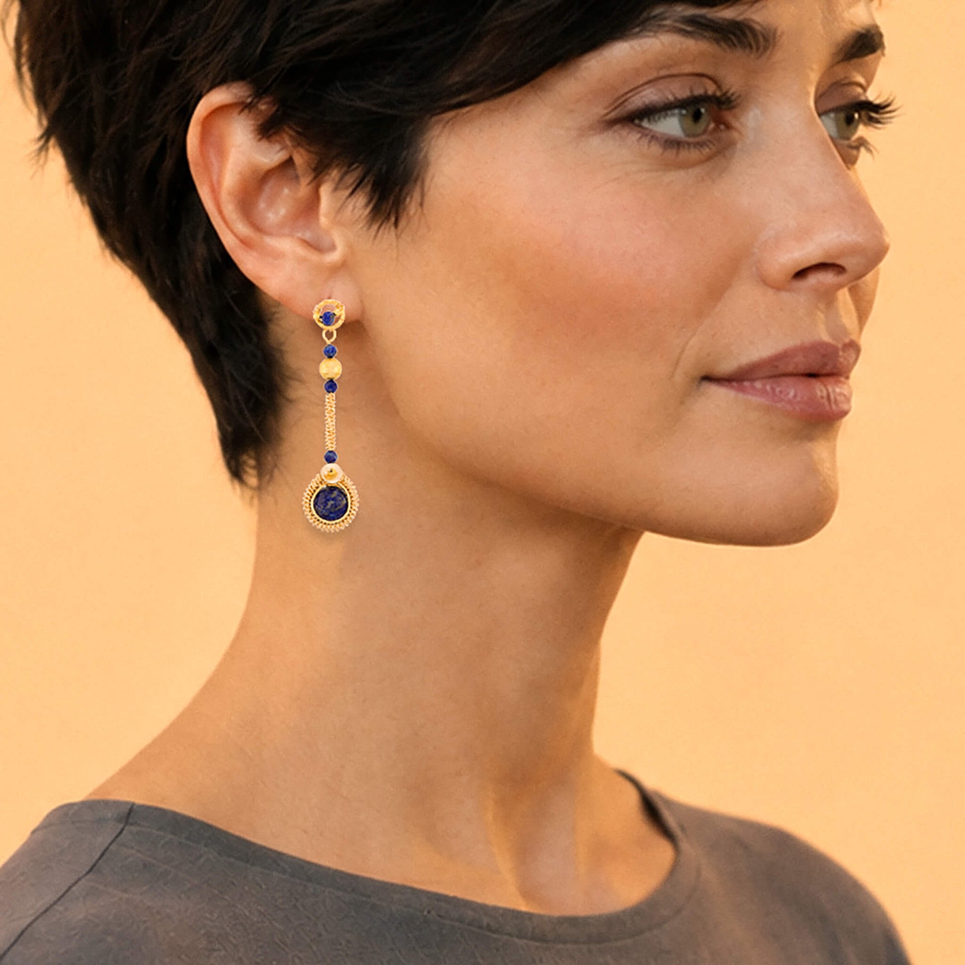 A close-up of a woman wearing a long gold dangle earring with deep blue circular stones and small gold and white accents, set against a warm beige background.