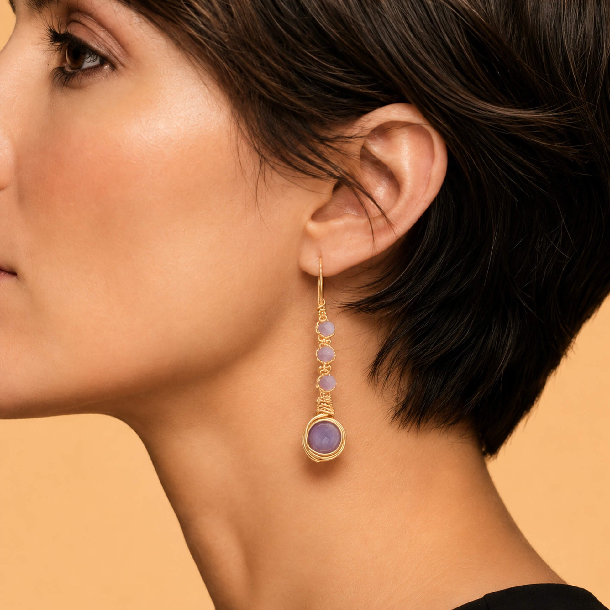 Close-up macro view of a gold wire-wrapped drop earring on a model's ear, showcasing the detailed wire craftsmanship and the texture of cascading purple amethyst stones.