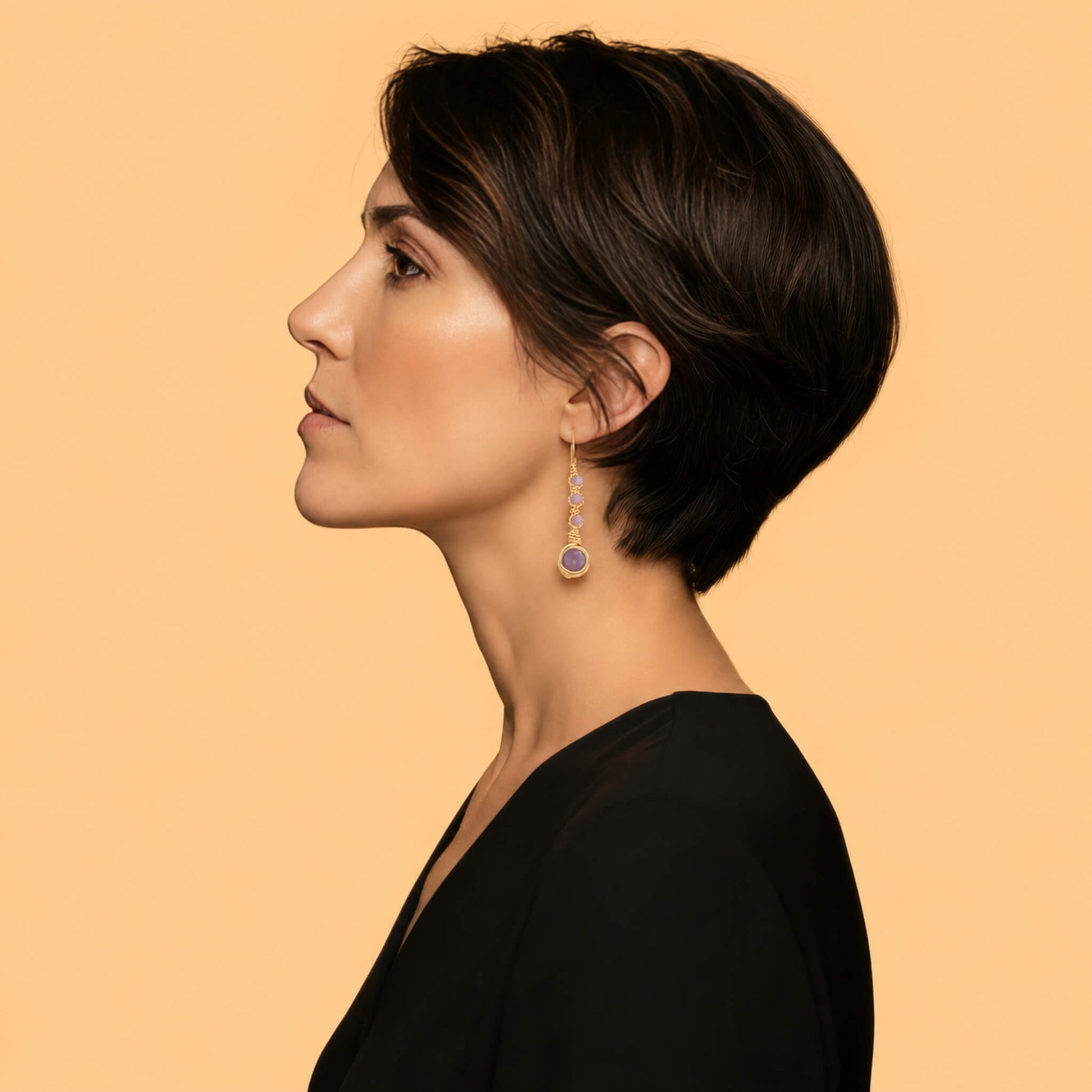 Lifestyle side profile of a woman with a short dark pixie cut wearing a gold and purple amethyst gemstone drop earring, styled with a minimalist black v-neck top against a warm beige background.