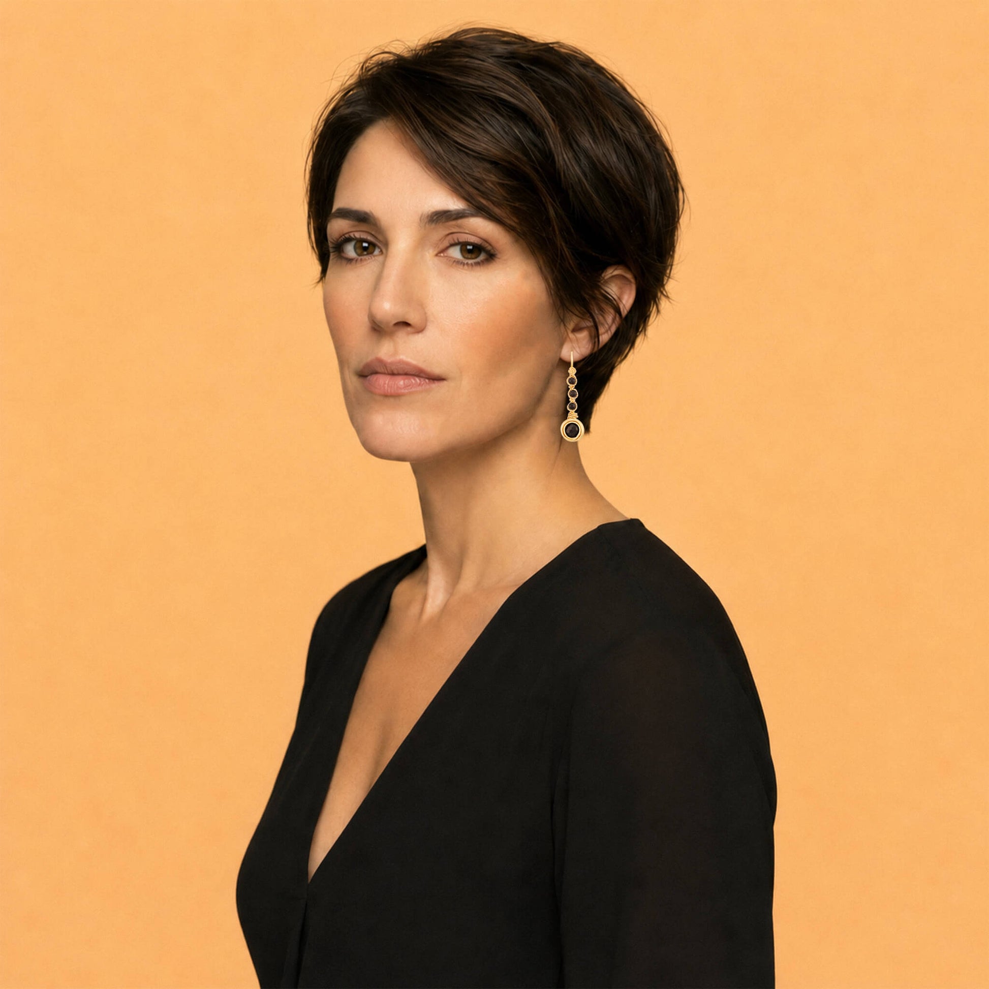 Profile lifestyle portrait of a woman with a dark pixie cut wearing a gold garnet long cascade earring, styled with a black V-neck top against a beige background.