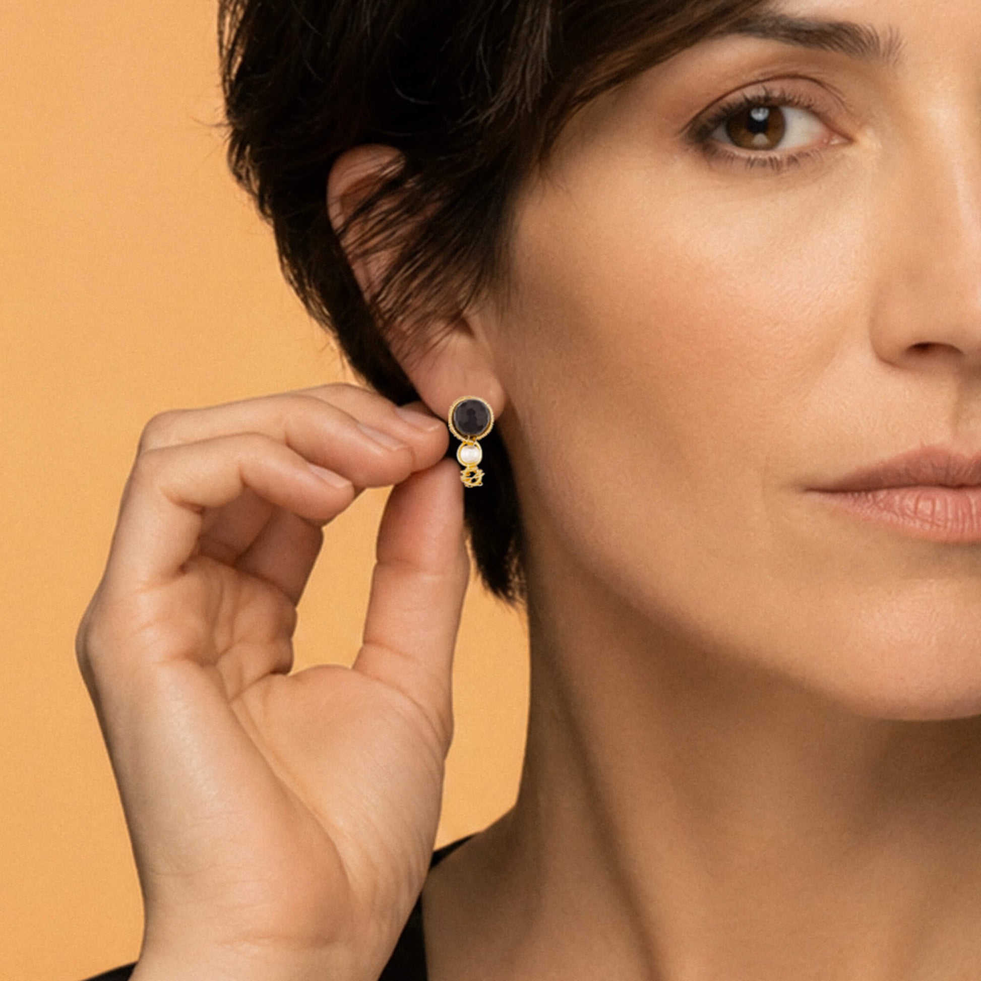 A close-up lifestyle shot of a model wearing a small gold hoop earring with a dark red Garnet gemstone, while holding the earring with her fingers to show its details against a warm beige background.