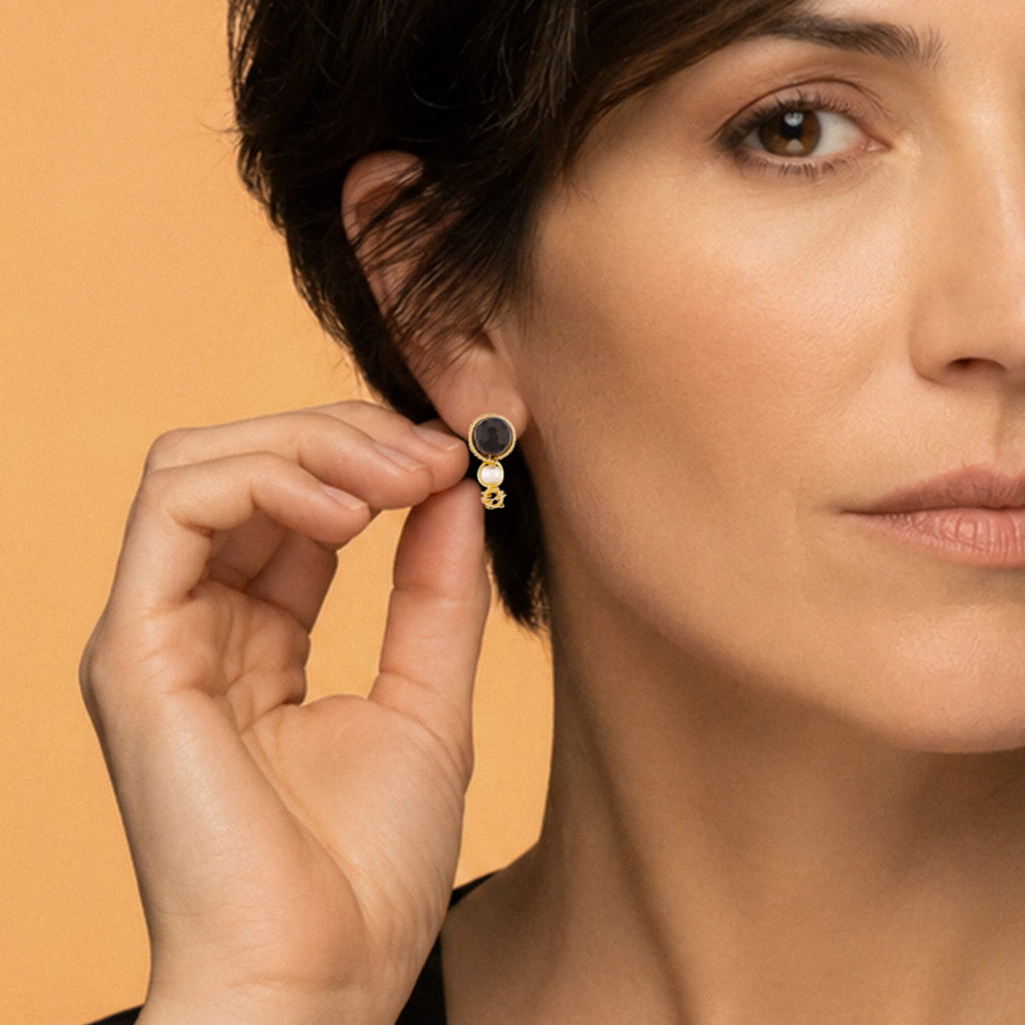 A close-up lifestyle shot of a model wearing a small gold hoop earring with a dark red Garnet gemstone, while holding the earring with her fingers to show its details against a warm beige background.