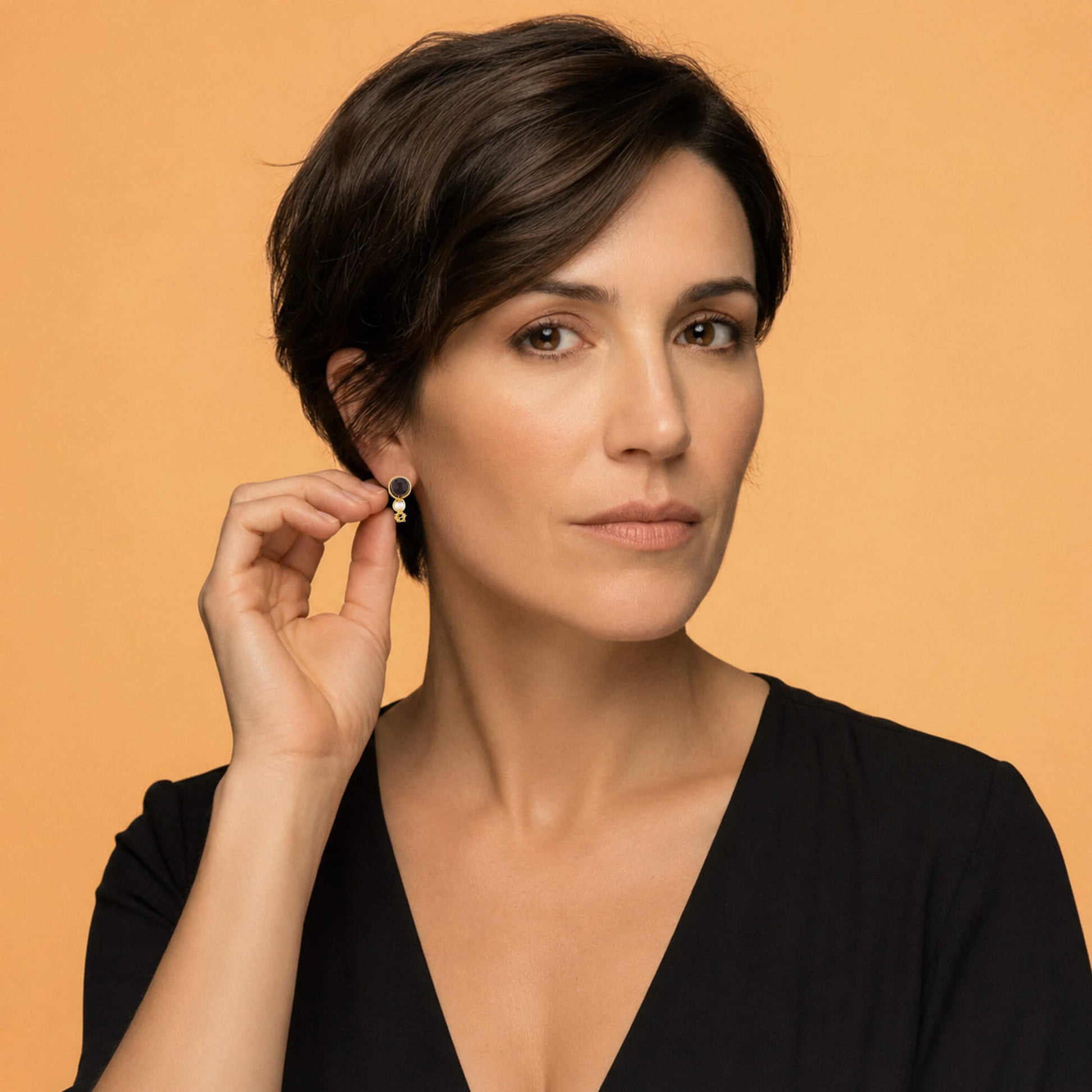 Portrait of a model with short dark hair wearing a small gold and dark red drop earring, holding it slightly with her hand against a solid warm orange background.