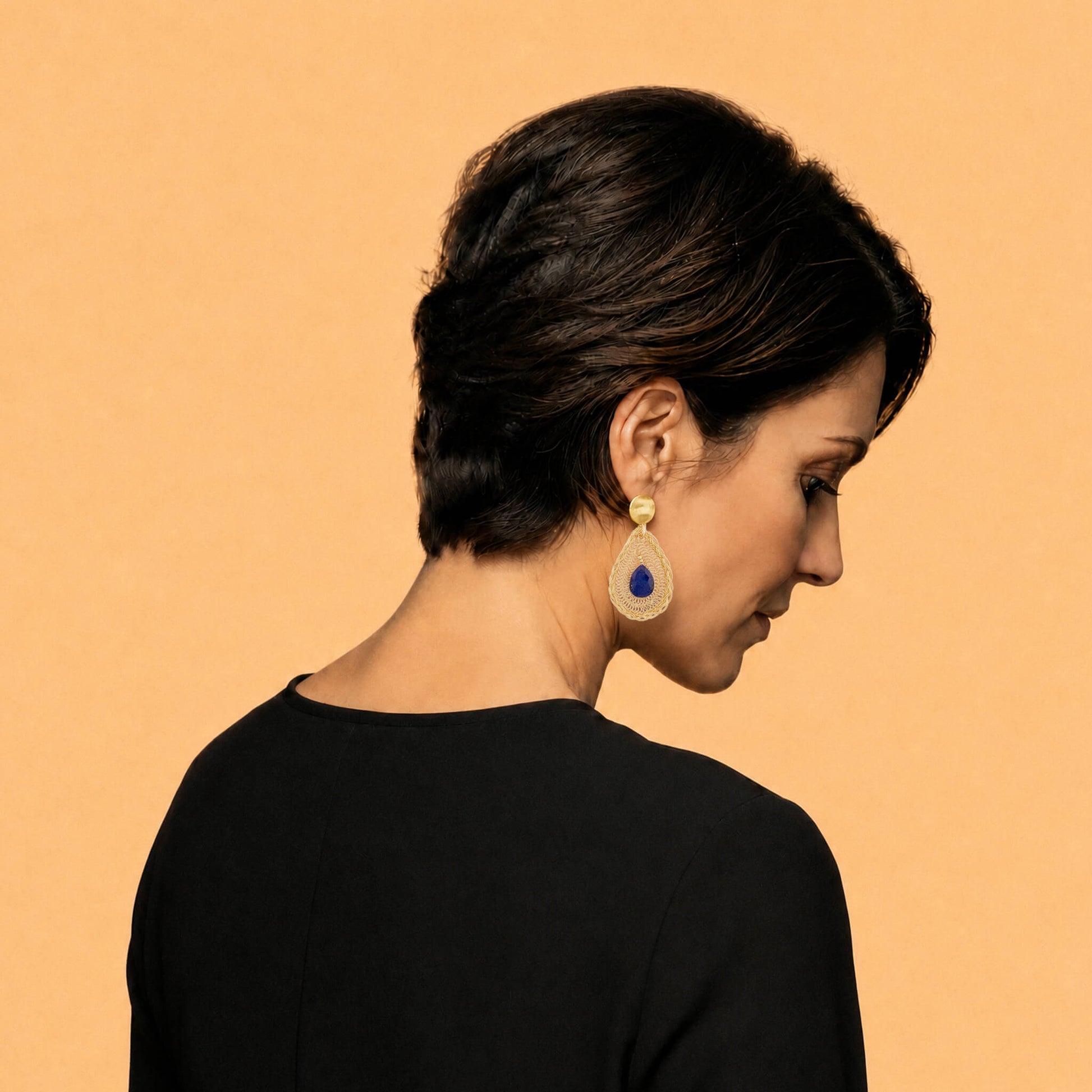 A model shown from the back and side profile wearing a single gold-toned teardrop earring with a deep blue Lapis Lazuli gemstone, set against a solid warm peach background.