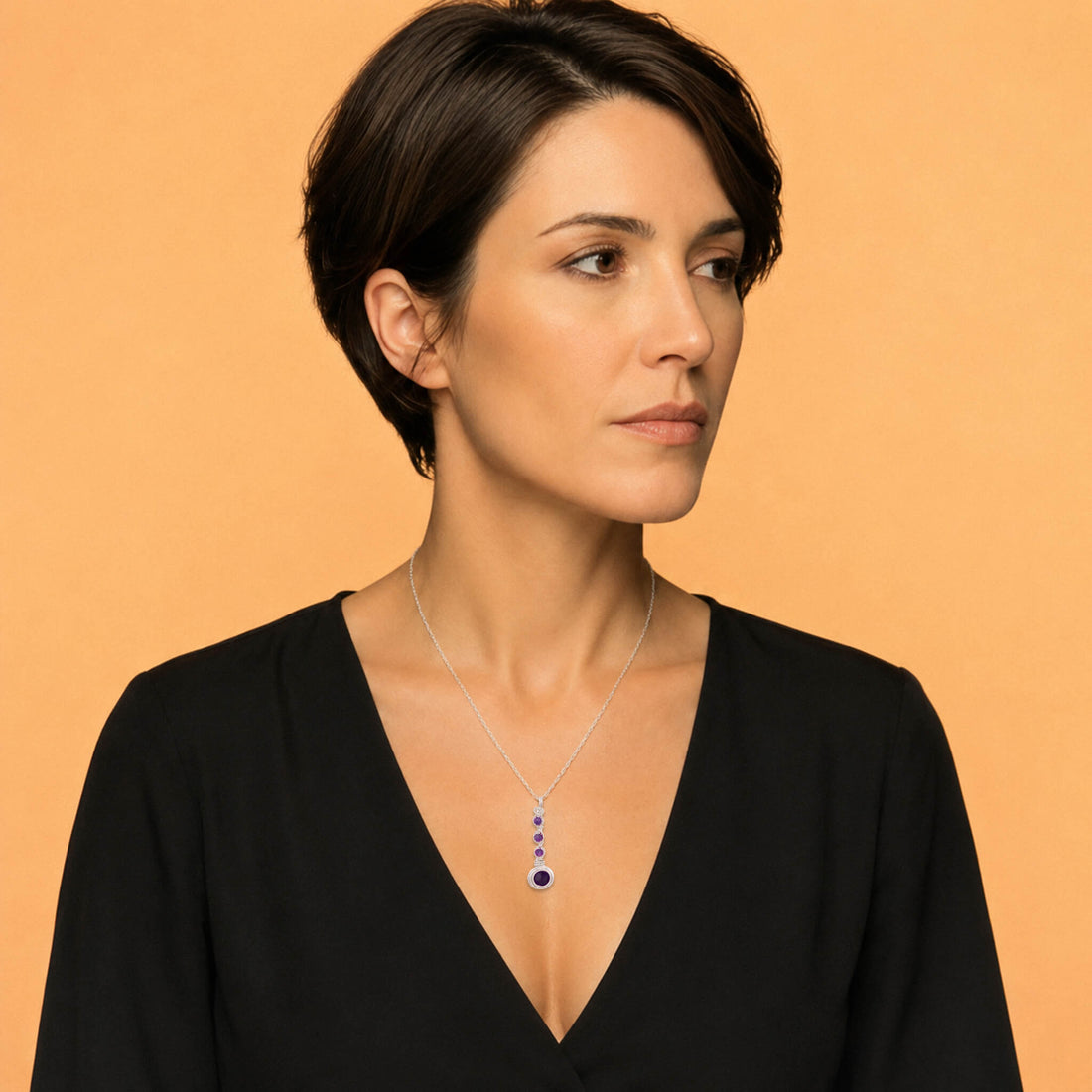 A silver paperclip chain necklace with a vertical drop pendant featuring three small round purple gemstones and a larger oval purple gemstone at the bottom, set against a plain white background.