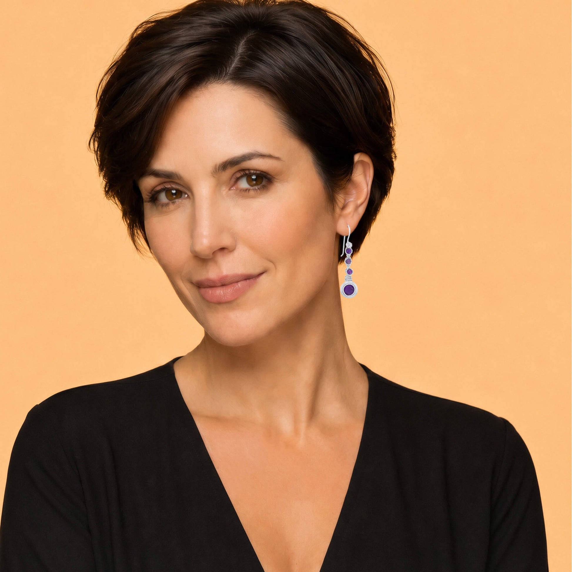 A woman with short dark hair wearing a black V-neck top and a dangling blue and silver earring, photographed against a solid warm peach background.
