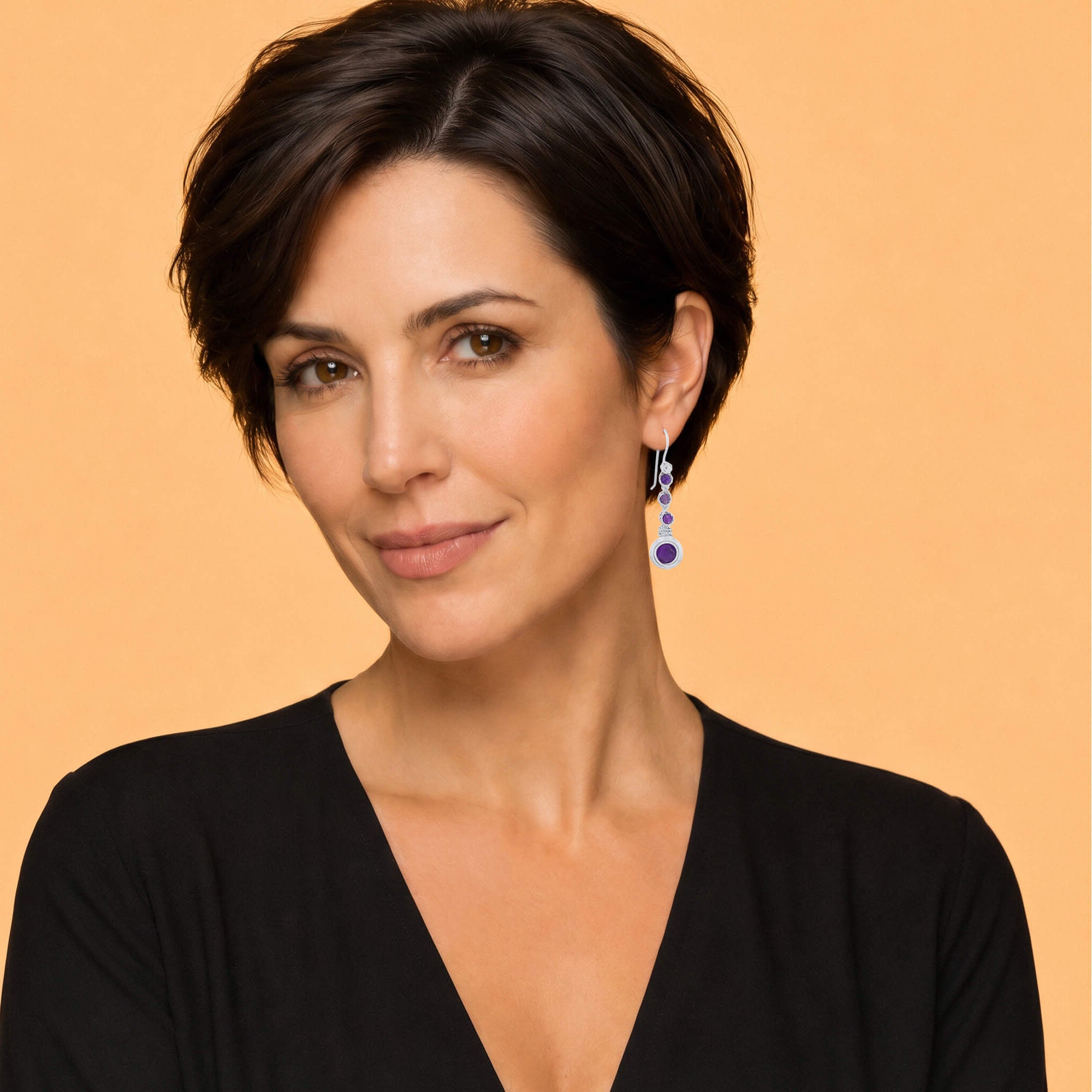 A woman with short dark hair wearing a black V-neck top and a dangling blue and silver earring, photographed against a solid warm peach background.