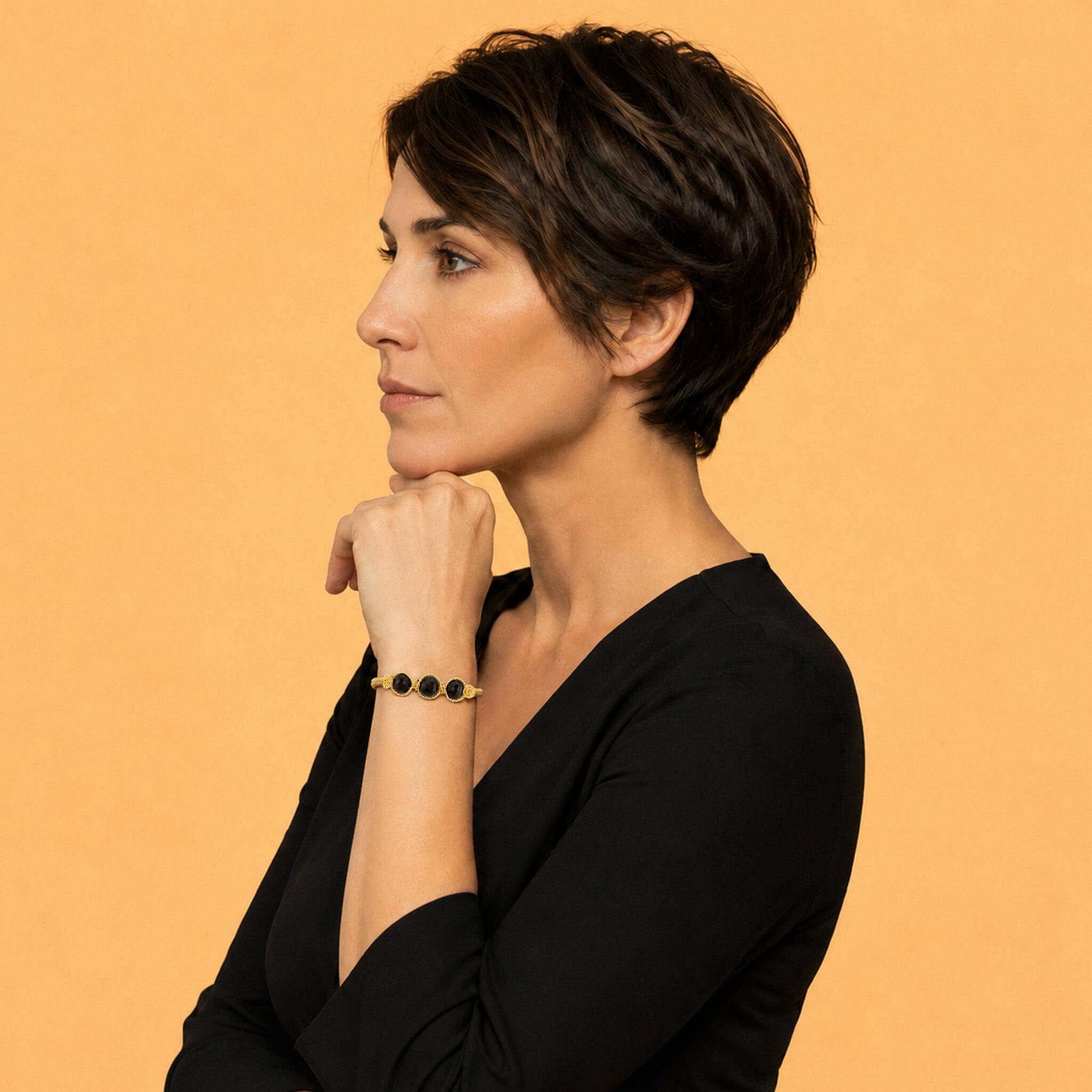A woman with short dark hair, wearing a black top and a gold bracelet with dark red garnet beads, posing in profile against a solid orange background.