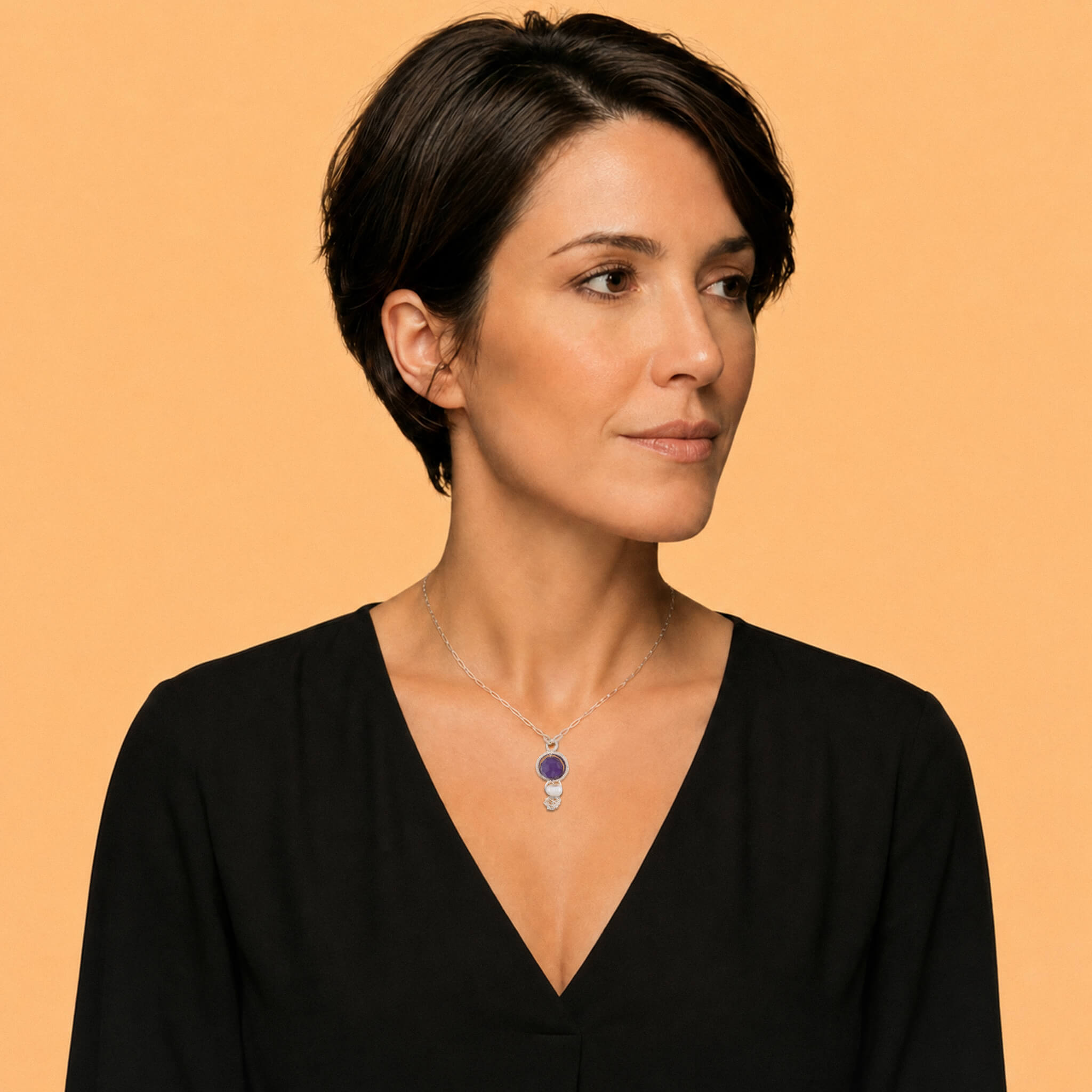 A woman with short dark hair wearing a black V-neck top and a silver pendant necklace with a purple round amethyst  gemstone, set against a solid warm beige background.