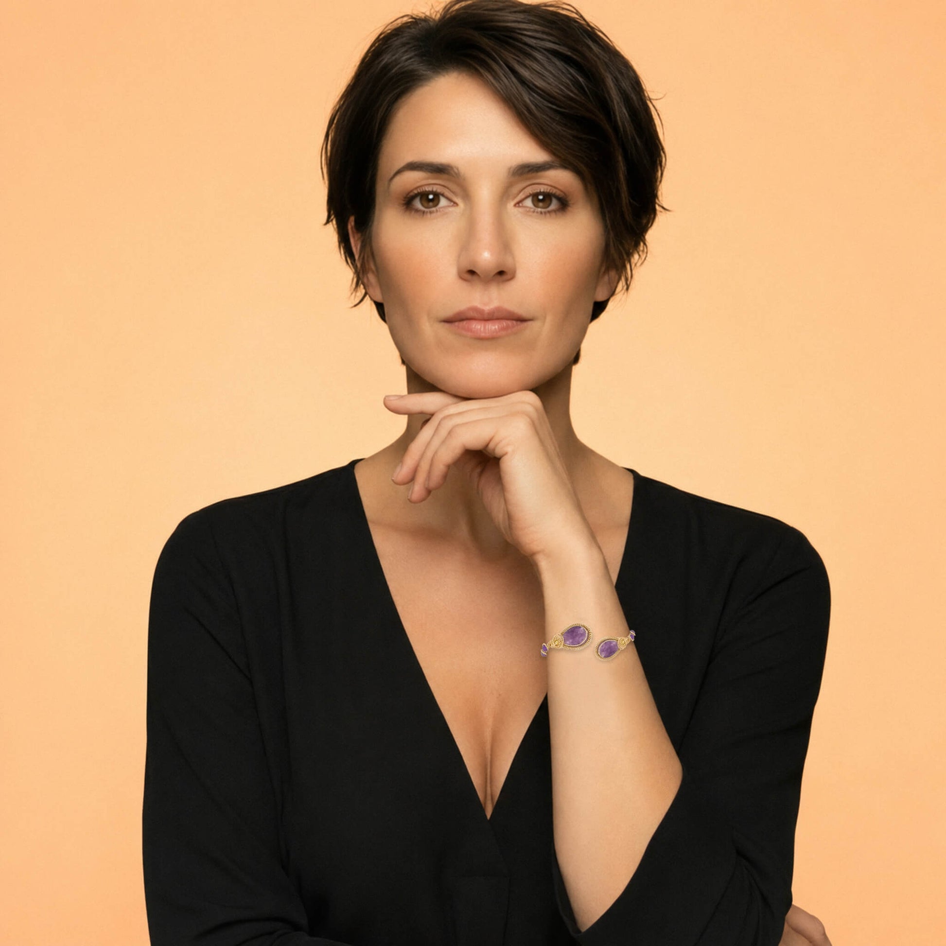 A woman with short dark hair wearing a black V-neck top and a delicate purple Amethyst bracelet, posing against a solid peach-colored background.