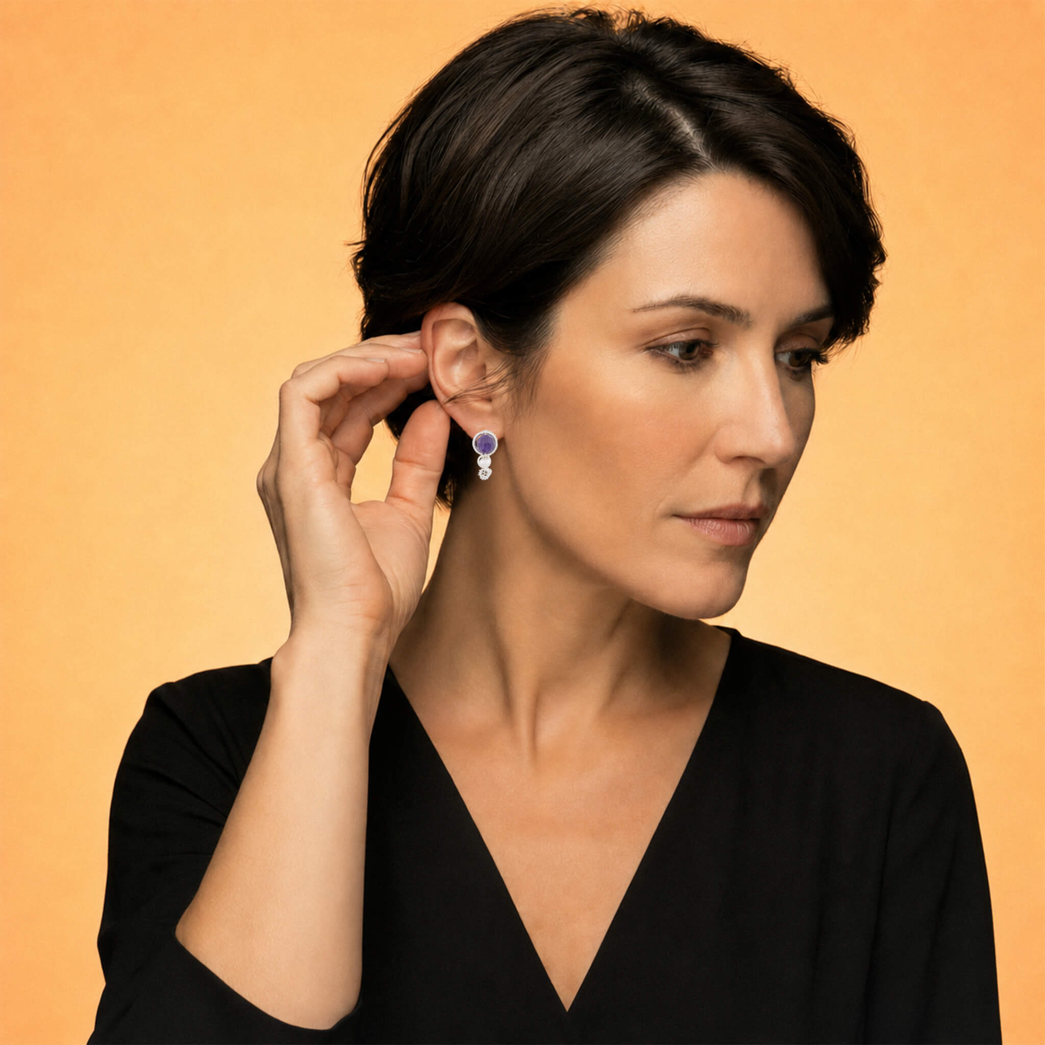 Woman with short dark hair displaying drop earrings round purple amethyst stone in silver bezel setting, white freshwater pearl, and small gold charm, worn against a warm amber background