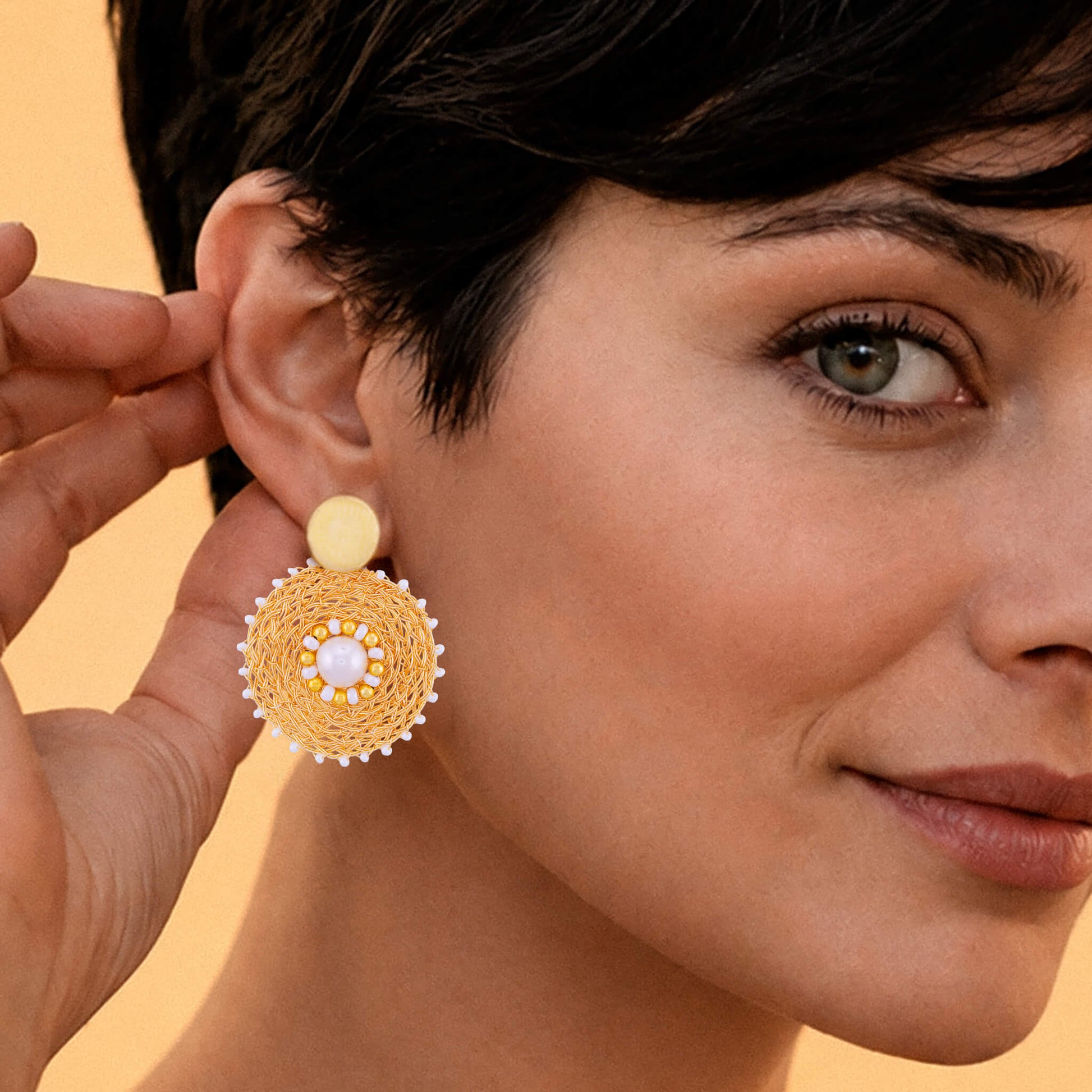 A close-up lifestyle shot of a woman wearing a large, circular gold-toned beaded earring with a white pearl-like center, holding the other side of the earring with her hand against a warm beige background.