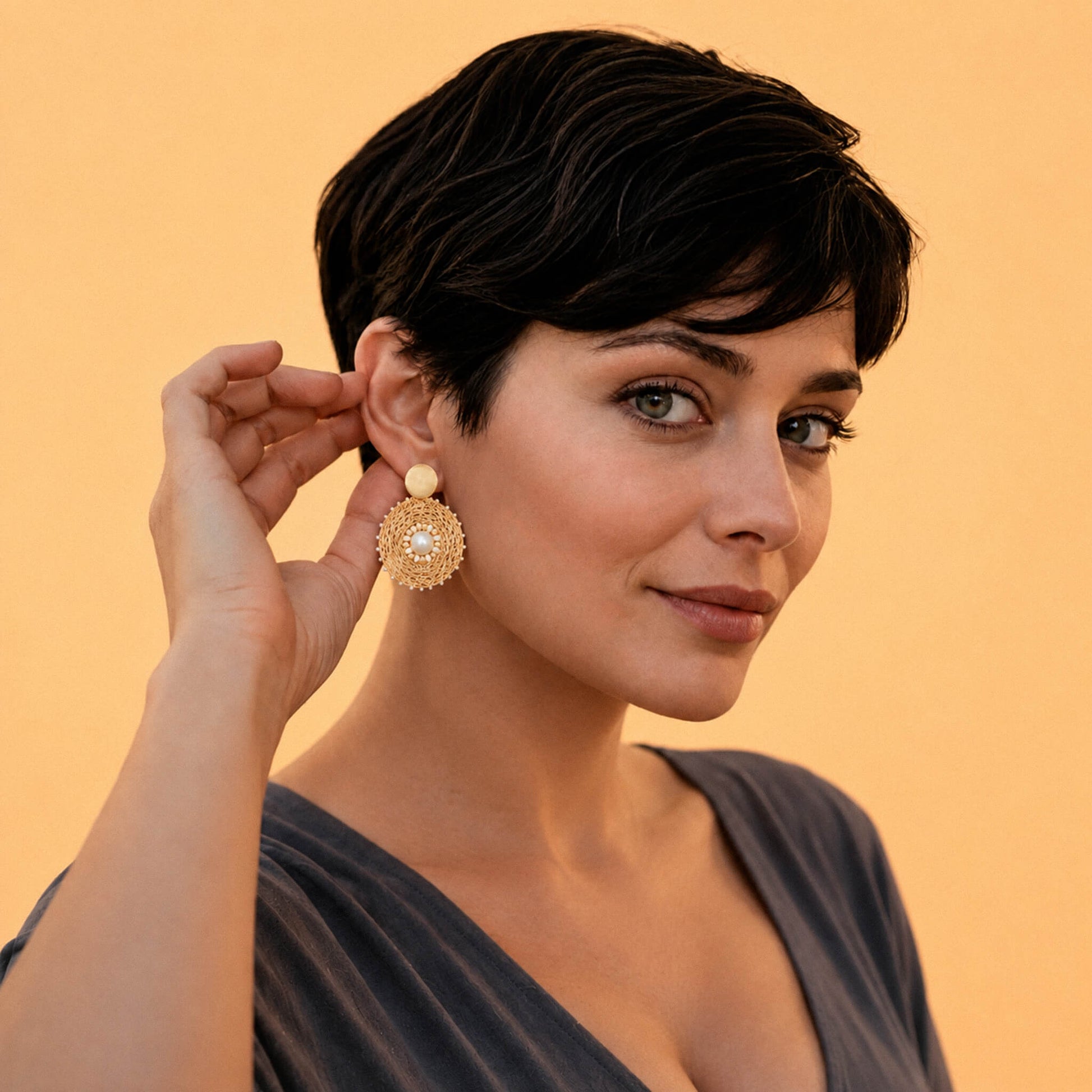 A woman with short dark hair models a large, ornate gold dangle earring with a cream-colored pearl-like top bead, set against a solid warm beige background.