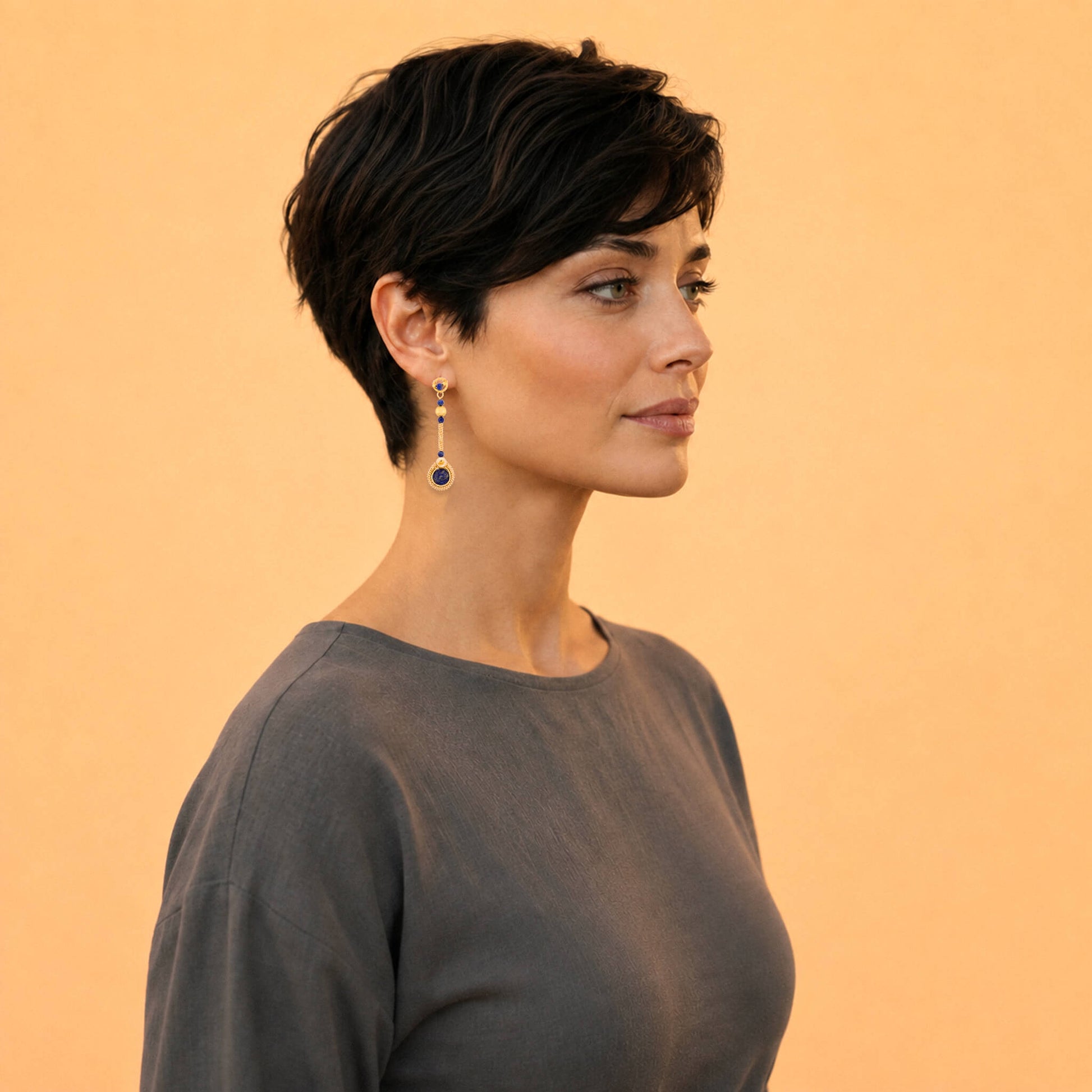 A woman with a short dark pixie cut is shown in profile against a solid warm peach background, wearing a dark gray top and a dangling drop earring with a gold-toned setting and a deep blue oval stone.
