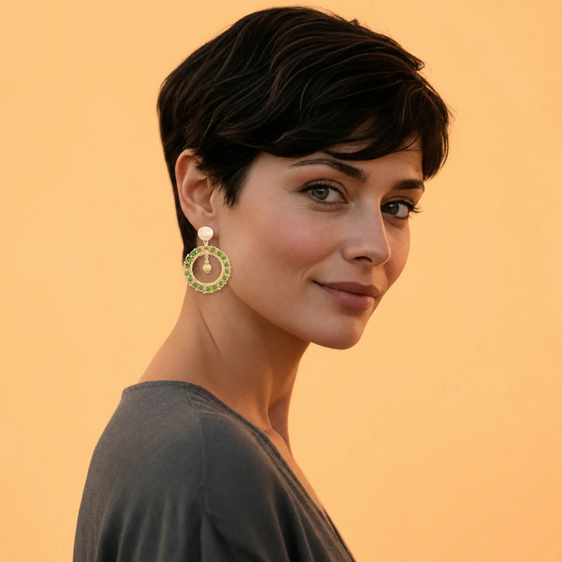 A pair of gold-toned statement dangle earrings featuring circular openwork wire frames accented with small green stones, a central gold bead drop, and a round textured gold post with a flat back.