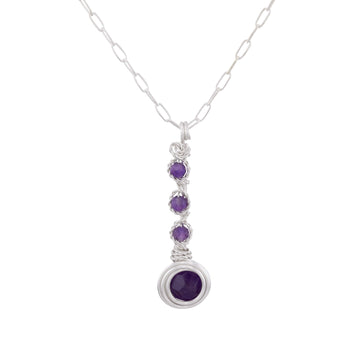 A silver paperclip chain necklace with a vertical drop pendant featuring three small round purple gemstones and a larger oval purple gemstone at the bottom, set against a plain white background.